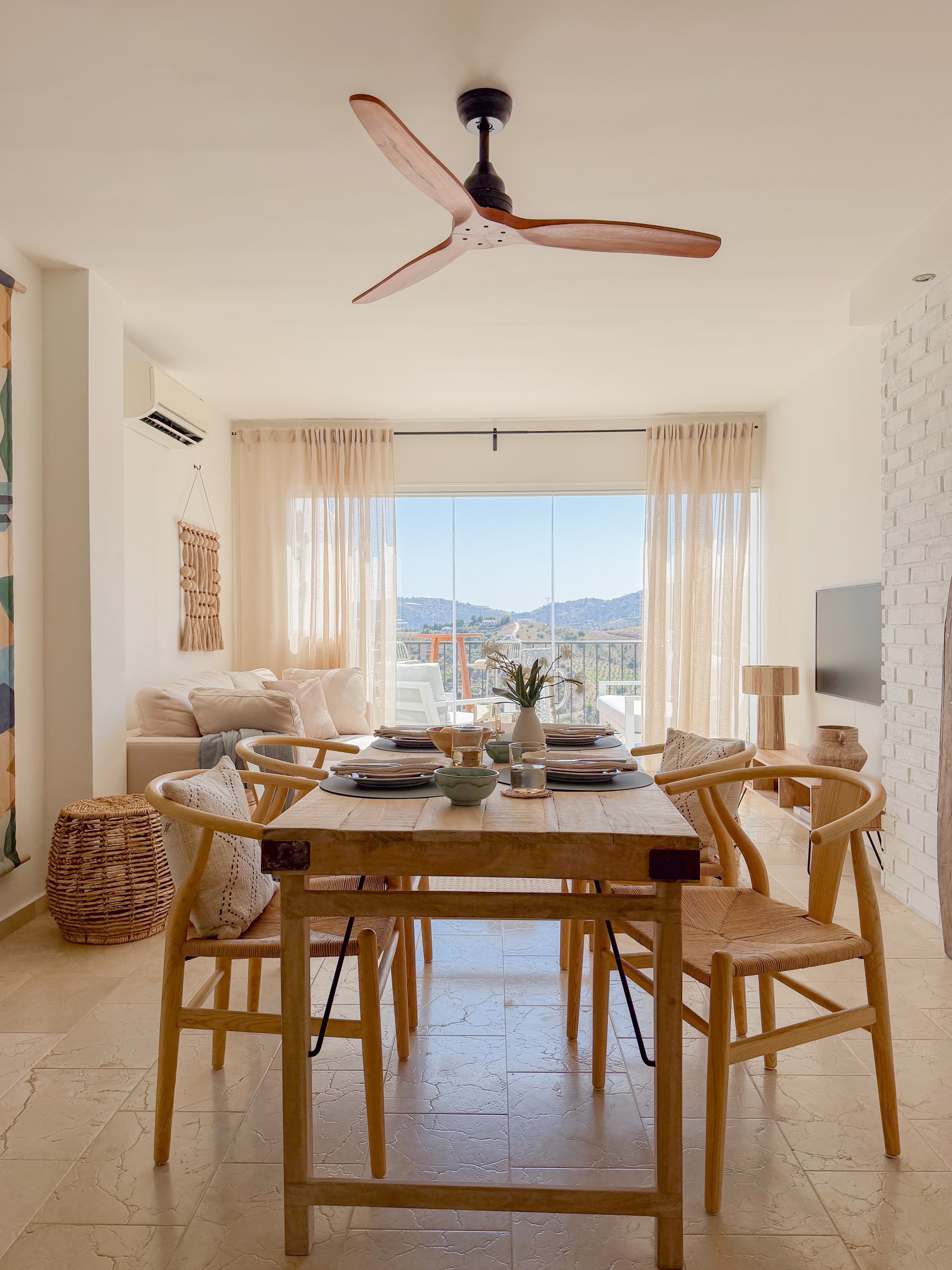Dining table set for a meal beneath a wood-bladed ceiling fan, with wishbone chairs and mountain views through the window