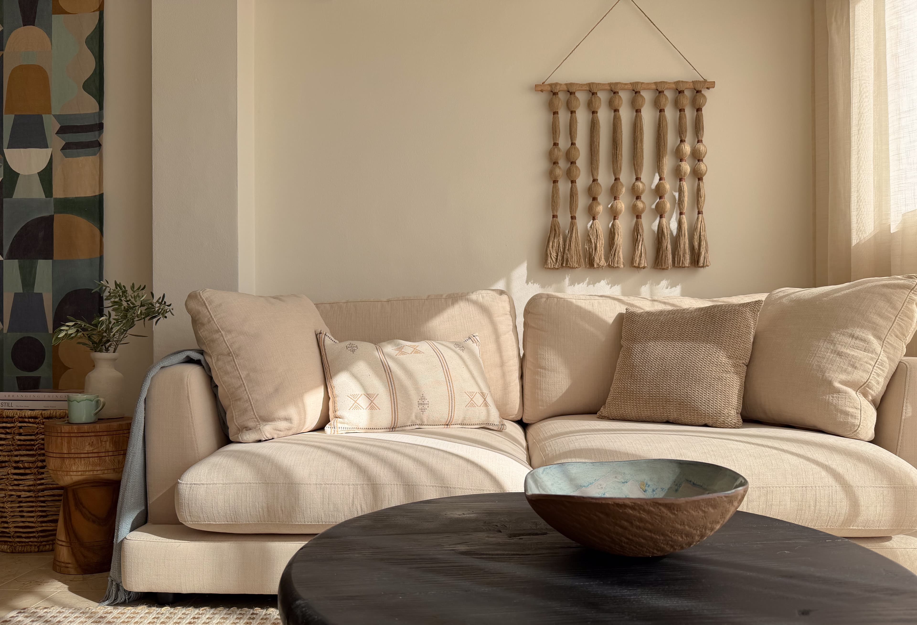 Living room sectional sofa beneath a tasseled wall hanging, with a colorful tapestry and dark coffee table