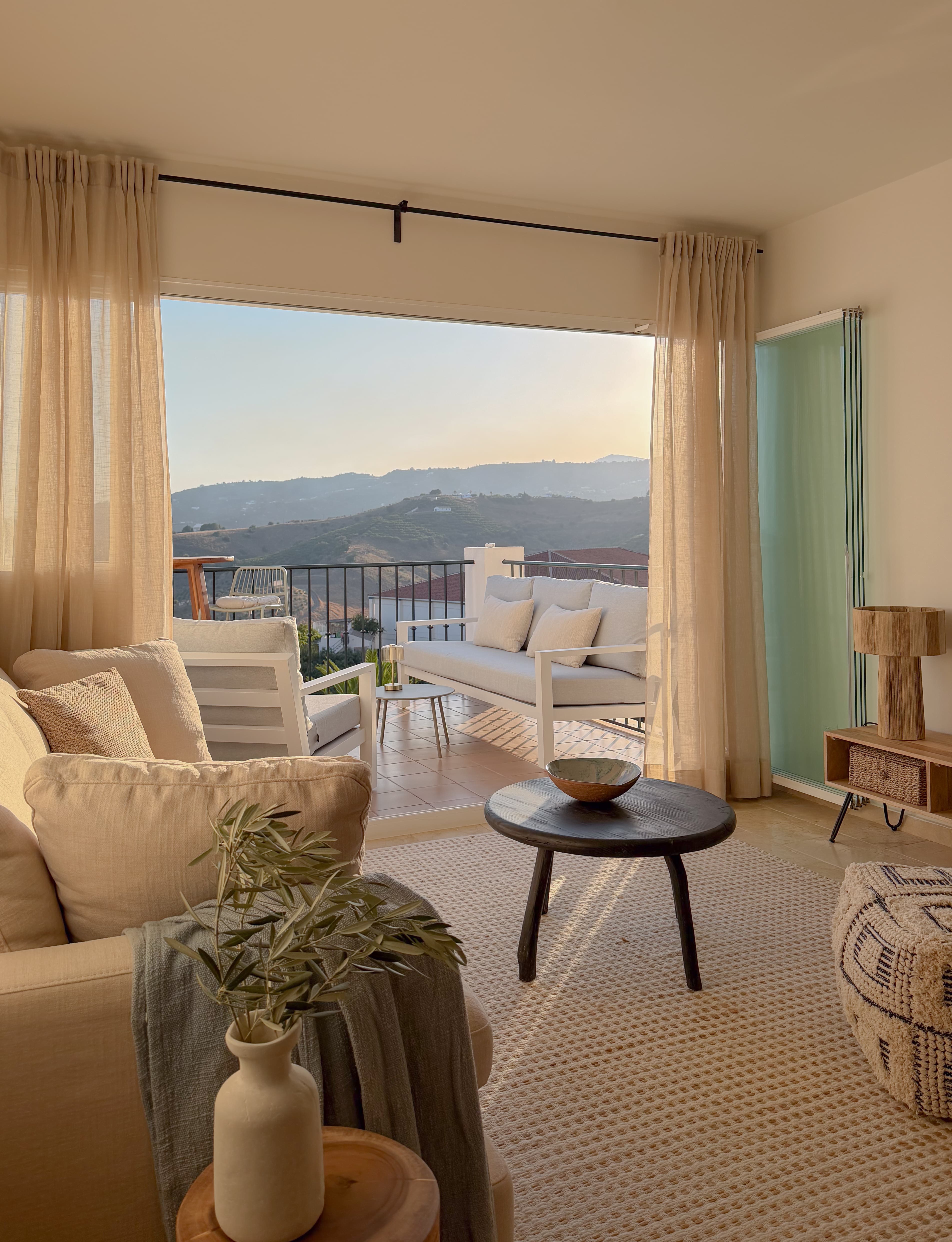Living room opening onto the terrace at golden hour, sofa and lounge chairs framed by sheer curtains with hillside views beyond