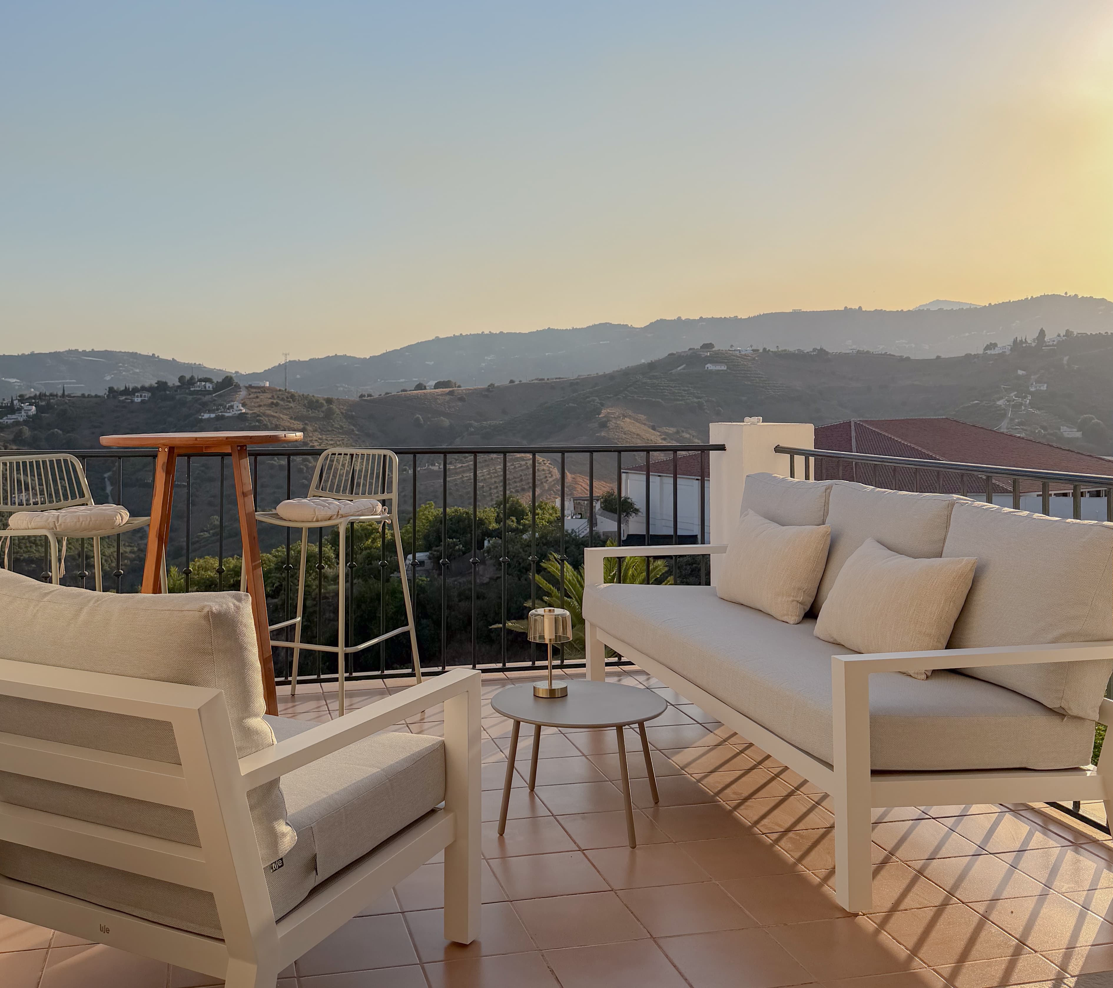Terrace lounge seating at sunset with a bar table and stools, overlooking hills and village rooftops