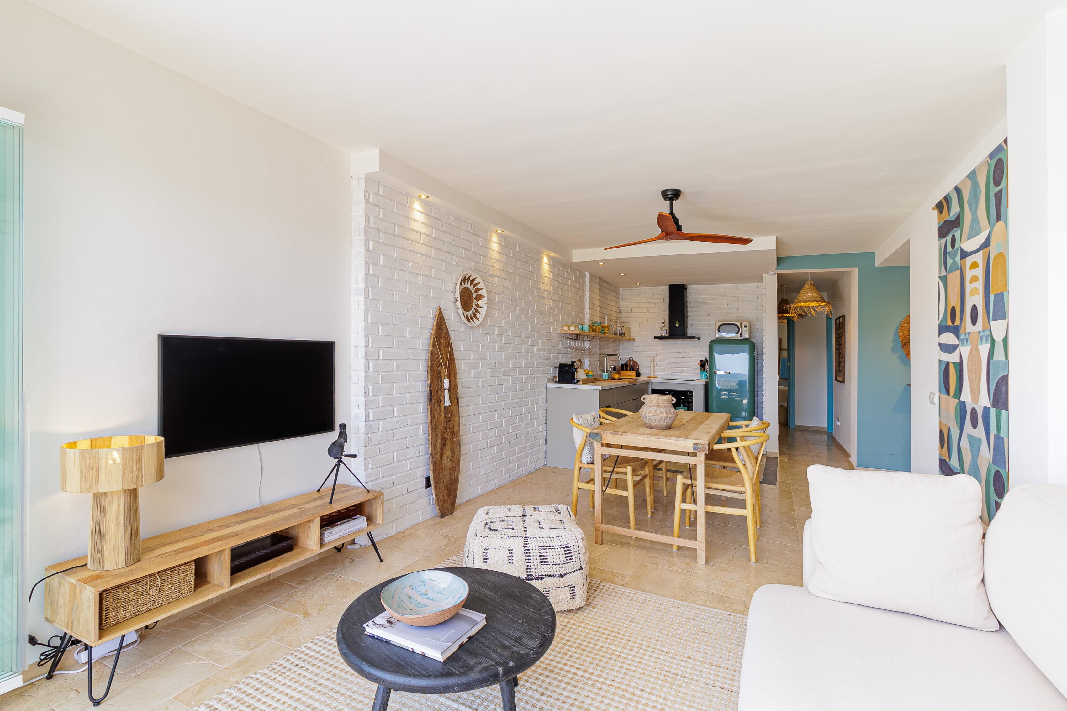 Living and dining area viewed from the TV corner, with a surfboard on the brick wall and a colorful geometric tapestry beyond